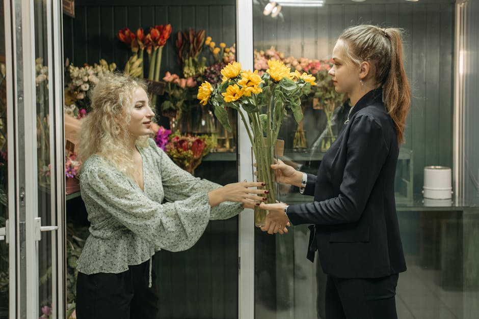 A woman with curly blonde hair, wearing a light gray blouse with a ruffled hem, is accepting a bouquet of bright yellow flowers with large petals and green stems from another woman with straight, redd