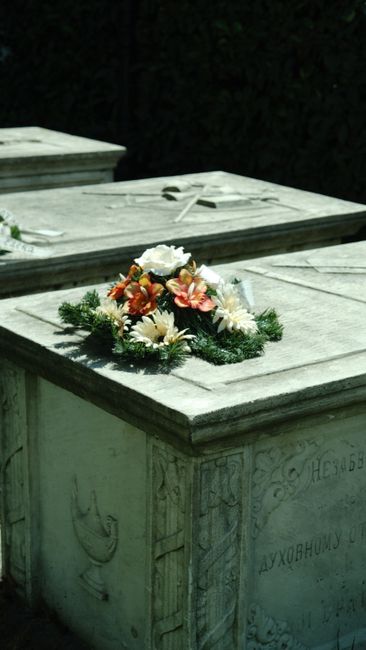 A floral arrangement placed on a stone grave marker, featuring a wreath of fresh flowers including white daisies, orange and pink lilies, and a white rose at the center, all encircled by green foliage