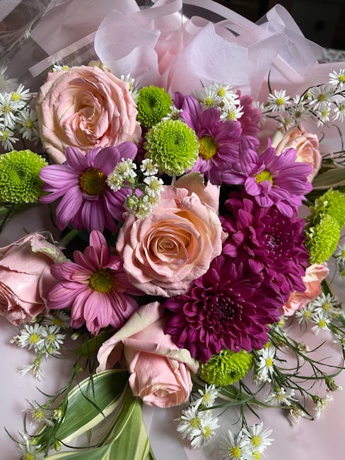 A vibrant floral arrangement featuring soft pink roses with tightly layered petals, surrounded by bright purple daisies with yellow centers, and lime-green chrysanthemums with dense, rounded blooms. S