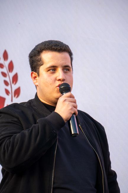 This image features a young man with short, dark curly hair speaking into a handheld microphone during an outdoor event. He is dressed in a black zip-up jacket over a black shirt, standing against a w